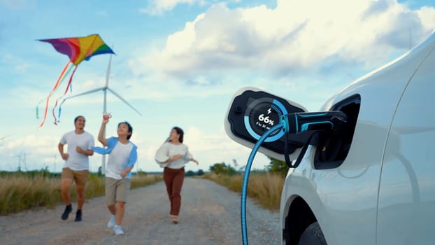 Focus eco-friendly EV car recharging display battery status hologram, charging station using eco-friendly energy wind turbine generator with happy family playing kite together in background. Peruse