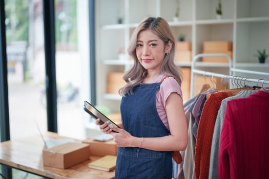 Portrait Of Asian Female Owner Of Fashion Store Checking Stock In Clothing Store With using notebook successful happy smile at small business, sme or ecommerce concepts.