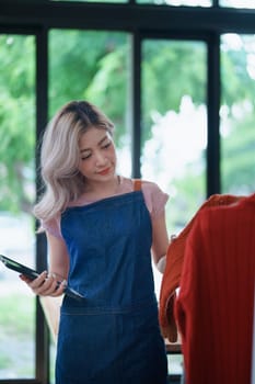 Portrait Of Asian Female Owner Of Fashion Store Checking Stock In Clothing Store With using notebook successful happy smile at small business, sme or ecommerce concepts.