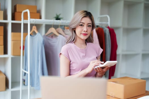 Portrait Of Asian Female Owner Of Fashion Store Checking Stock In Clothing Store With using notebook successful happy smile at small business, sme or ecommerce concepts.