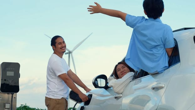 Concept of progressive happy family enjoying their time at wind farm with electric vehicle. Electric vehicle driven by clean renewable energy from wind turbine generator for charging station.