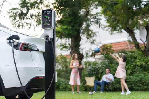 Focus electric vehicle recharge from home charging station on blur background of happy and playful family playing together. EV car using alternative and sustainable energy for better future.Synchronos