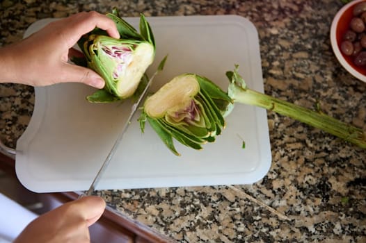 View from above of chef hands cutting artichoke flower on a board. Two halves of fresh ripe organic artichoke on cutting board