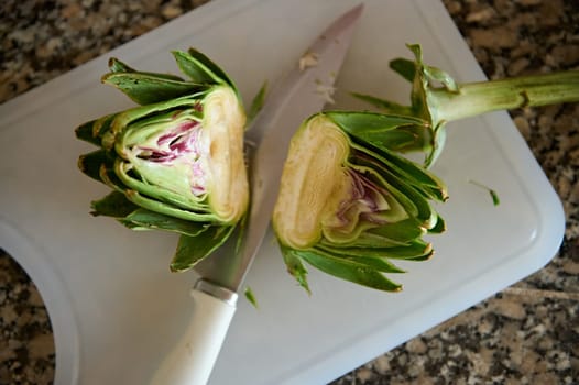 Two halves of fresh ripe organic artichoke and a kitchen knife on cutting board, on marble stone kitchen counter. Raw vegan food background. Healthy eating, m dieting concept