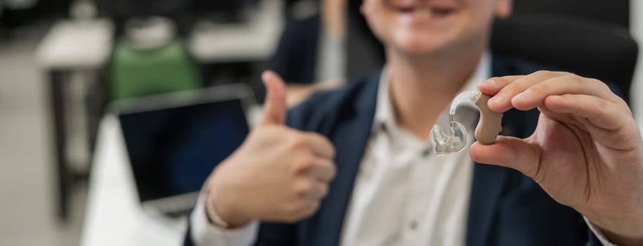Portrait of a man holding a hearing aid and showing thumbs up in the office. Widescreen