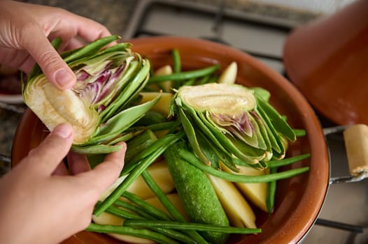 Closeup top view of woman's hands putting halves of artichoke flower on a clay dish with vegetables, cooking tagine at home. Traditional Moroccan cuisine, culinary, Culture and traditions