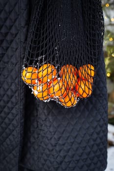 The girl is holding bright orange tangerines in a black string bag in her hands. It's snowing outside