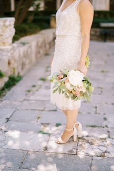 Bride with a bouquet of flowers stands half-turned on the paving stones. Cropped. Faceless. High quality photo