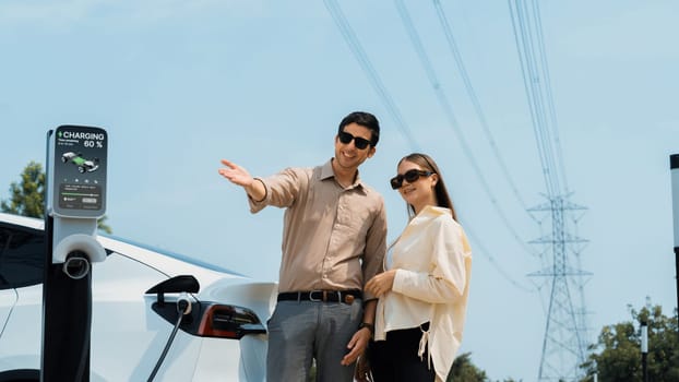 Young couple recharge EV car battery at charging station connected to power grid tower electrical industrial facility as electrical industry for eco friendly vehicle utilization. Expedient