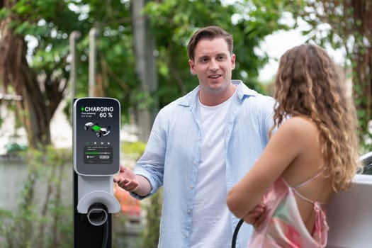 Happy and lovely couple with eco-friendly conscious recharging electric vehicle from EV charging station. EV car technology utilized as alternative transportation for future sustainability. Synchronos