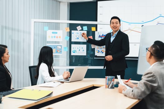 Successful businessmen present financial projects on glass whiteboard confidently. Executive managers listen to employee's quarterly business plan presentation. Modern meeting room. Intellectual.