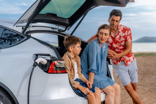 Family vacation trip traveling by the beach with electric car, lovely family sit on the trunk, charging EV car battery with green and sustainable energy. Family travel and eco-friendly car. Perpetual