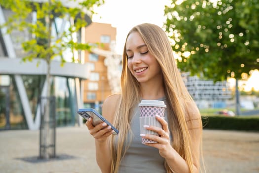 Attractive blonde hair woman uses her smartphone and drinks coffee in the street.