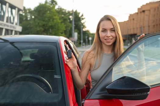 Female with long blonde hair standing with key near new red car in the street.