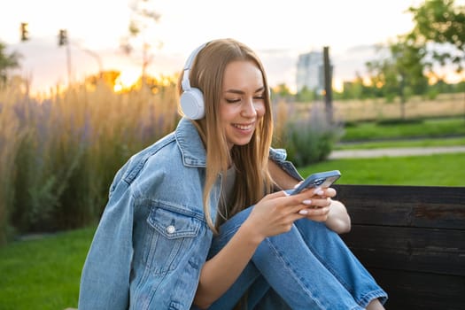 Attractive young woman, wearing a denim, relaxing in garden and listening to music trough headphones and smart phone