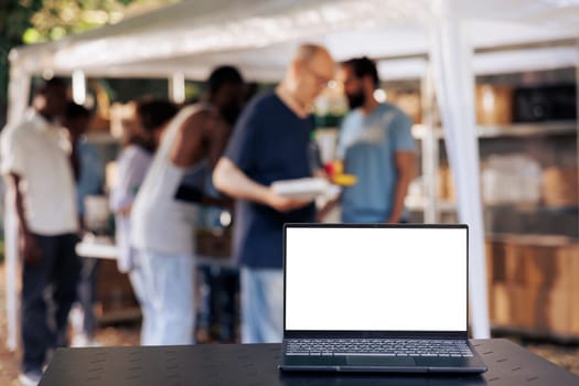 Image showcasing laptop displaying isolated white screen placed on table while group of volunteers provide assistance to the needy. Photo focus on minicomputer with a blank copyspace mockup template.