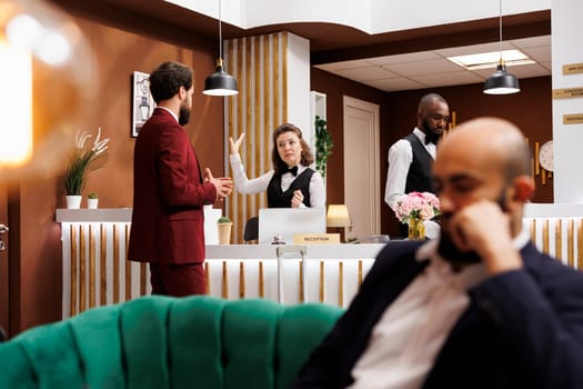 Businessman chats with front desk staff in reception lobby, asking about hotel room services after doing check in. Formal guest arriving at resort, travelling on business trip.