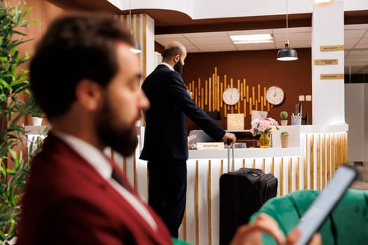 Businessman with luggage at front desk waiting to see room reservation and check in, travelling for work. Company professional preparing for upcoming business conferences, hotel lobby.