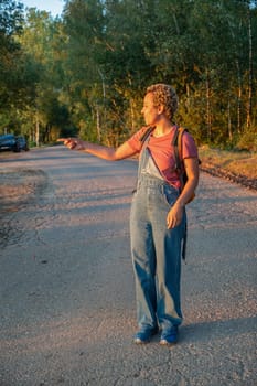 Brazilian millennial girl in denim overalls stands on a forest road and points with her hand at sunset, golden hour on the lake, High quality photo