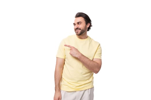 young brutal brunette man in a T-shirt points his hand to the side on a white background with copy space.