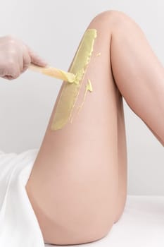 Hand in glove applying green hot wax on slim woman leg using spatula. Depilation procedure with hot wax in beauty salon. Part of photo series.
