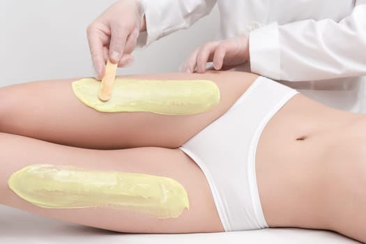 Beautician in gloves applying hot wax on slim woman legs using spatula while woman lying down on couch. Depilation with hot wax in beauty salon. Healthy body in white panties. Part of photo series.