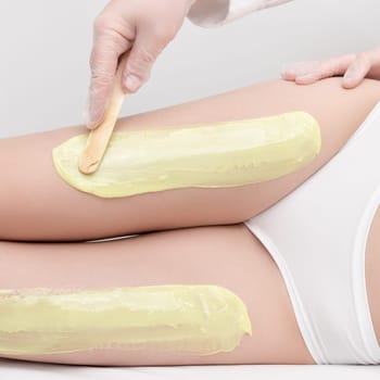 Cosmetologist in gloves applies green hot wax on slim woman legs using spatula while woman lying down on couch. Depilation with hot wax in beauty salon. Close up side view. Part of photo series.