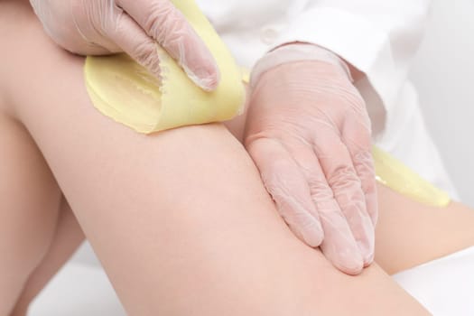 Closeup of cosmetologist hands doing depilation to woman legs with green hot wax in beauty salon. Waxing process of hair removal from root. Women intimate hygiene concepts. Part of photo series.