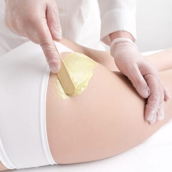 Closeup hand applying hot wax on woman buttocks using spatula during hair removal procedure. Waxing process, depilation with green hot wax in professional beauty salon. Part of photo series