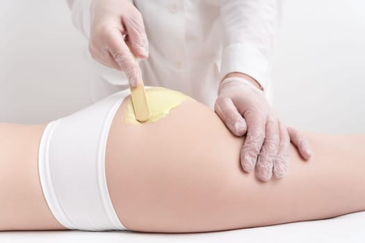 Unrecognizable beautician in gloves applying green hot wax on buttocks of slim woman using spatula. Waxing procedure, depilation process with hot wax in professional beauty salon. Part of photo series