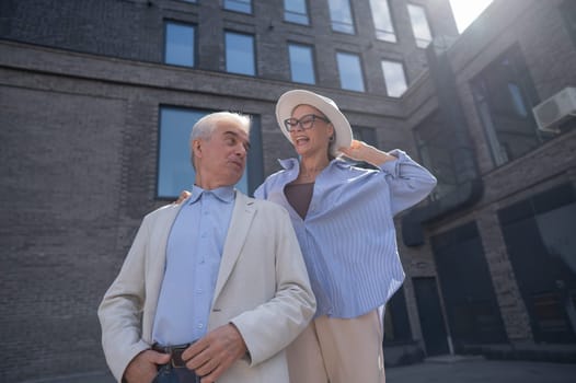 A woman in a hat and glasses hugs an elderly man in a white jacket from behind. Romantic relationships of mature people