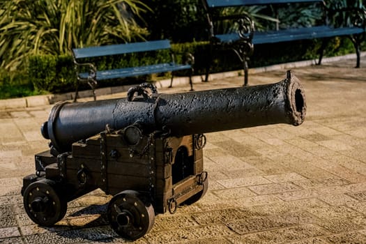 Old bronze cannon on a wooden cart with wheels stands on the paving stones near the bench. High quality photo