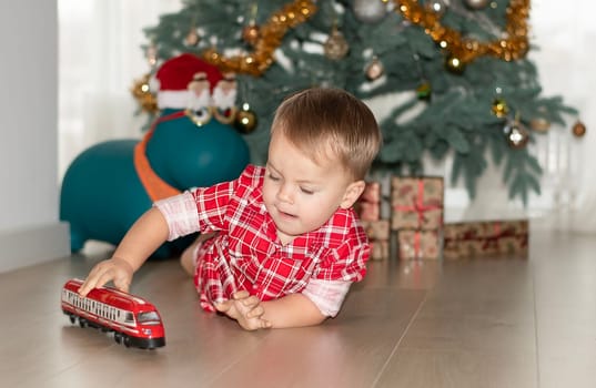 Christmas concept. New Year 2024. A small handsome boy in red checkered pajamas lies on the floor and plays with a red steam locomotive against the background of a decorated Christmas tree with gifts. Close-up.