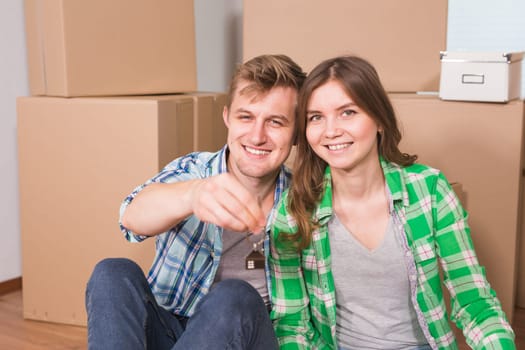 Happy young couple sitting on floor near boxes. Young family moving to new home. Woman and man showing keys and smiling at camera.