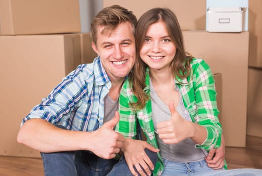 Moving to a new house and repairs in the apartment. Love couple showing a thumbs up and sitting in an empty apartment among boxes.