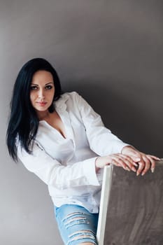 brunette woman in a white shirt sits on a chair on a gray background