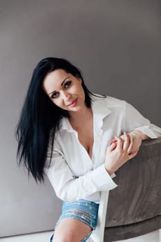 brunette woman in a white shirt sits on a chair on a gray background