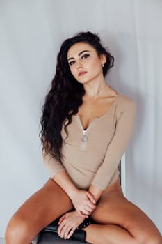 brunette woman in a bodysuit sits against a white wall
