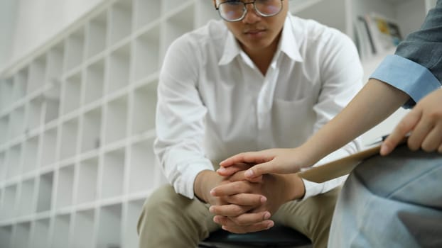Psychiatrist or counselor holding the patient's hand to comfort and encourage. Young Asian female psychologist keeping hand of wrist of male patient sitting in front of her and sharing his problems.