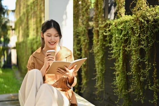 Happy young Asian woman drinking coffee and reading book in garden.