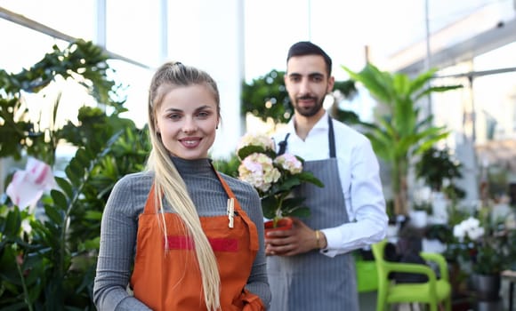 Smiling Woman Florist and Male with Hydrangea. Blonde Girl Gardener in Apron and Man Holding White Flower Hortensia in Pot. People Workers at Domestic Plant Center Looking at Camera Photography