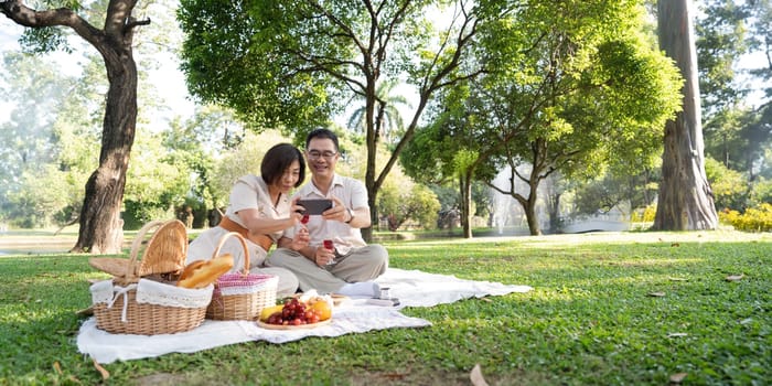 Cheerful elderly couple asian wear casual clothes sitting in the park having a party and taking selfie together.