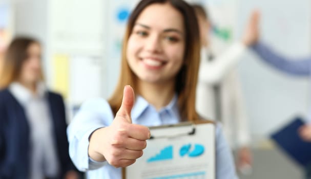 Focus on tender woman hand showing approving gesture at camera. Smiling lady holdig paper tablet with important document and standing in big modern office. Company meeting concept. Blurred background