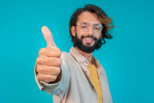 Young man doing happy thumbs up gesture with hand standing over isolated blue background. Approving expression looking at the camera showing success. High quality photo
