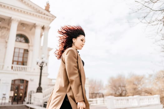 Woman street lifestyle. Image of stylish woman walking through European city on sunny day. Pretty woman with dark flowing hair, dressed in a beige raincoat and black, walks along the building