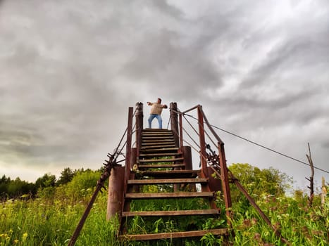 High, old, rusty iron staircase and tiny statue of girl or woman at the top against backdrop of gray, gloomy, thunderous clouds. Concept of an unusual, fantastic journey, surrealism, strange world