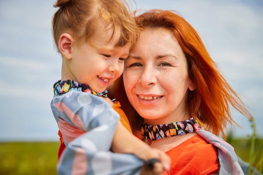 Happy female family with mother and daughter on green and yellow meadow full of grass and flower. Woman with red hair and blonde girl having fun, joy and hug in sunny summer day. Concept family love