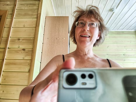 A shaggy, funny middle-aged woman in glasses takes selfie on her smartphone in front of the mirror in the morning. A blogger girl with disheveled hair takes pictures of herself for a blog