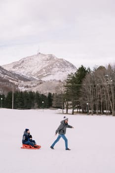 Dad carries mom and child on a sleigh across a snowy plain at the foot of the mountains. High quality photo