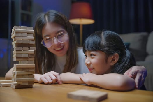 Asian young mother playing game in wood block with her little daughter in home living room at night time, Smiling woman help teach child play build constructor of wooden blocks, education